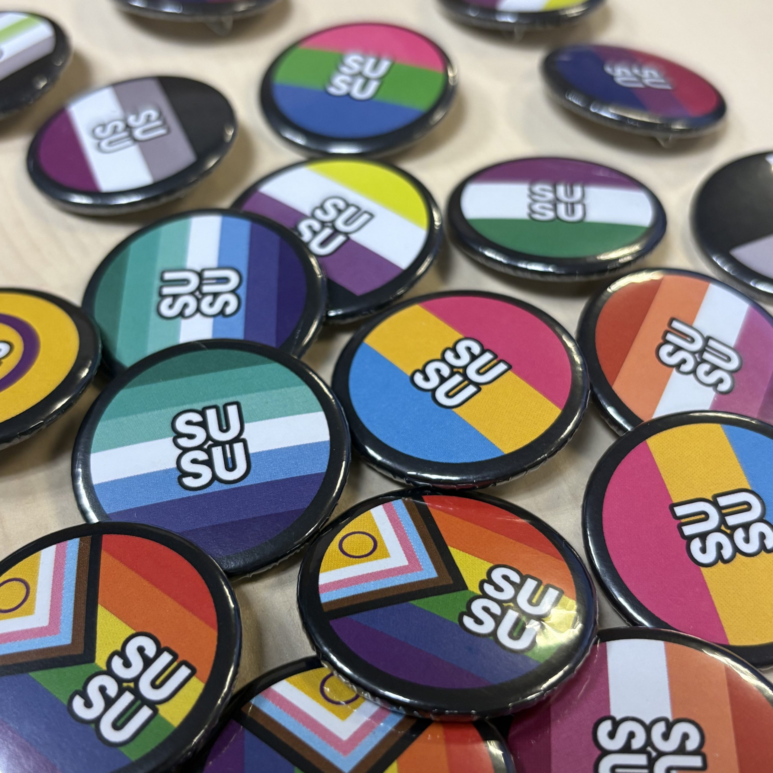A collection of SUSU-branded badges laid out on a table, each featuring different LGBTQ+ pride flag colours with the SUSU logo in the centre.